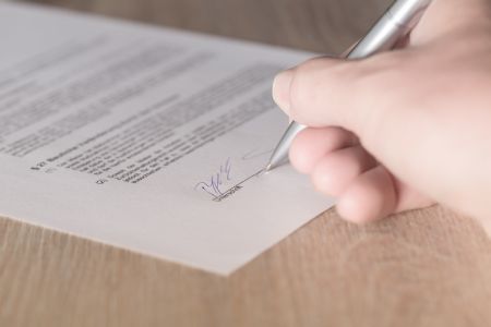 A Caucasian hand sighing a document on a wooden table.